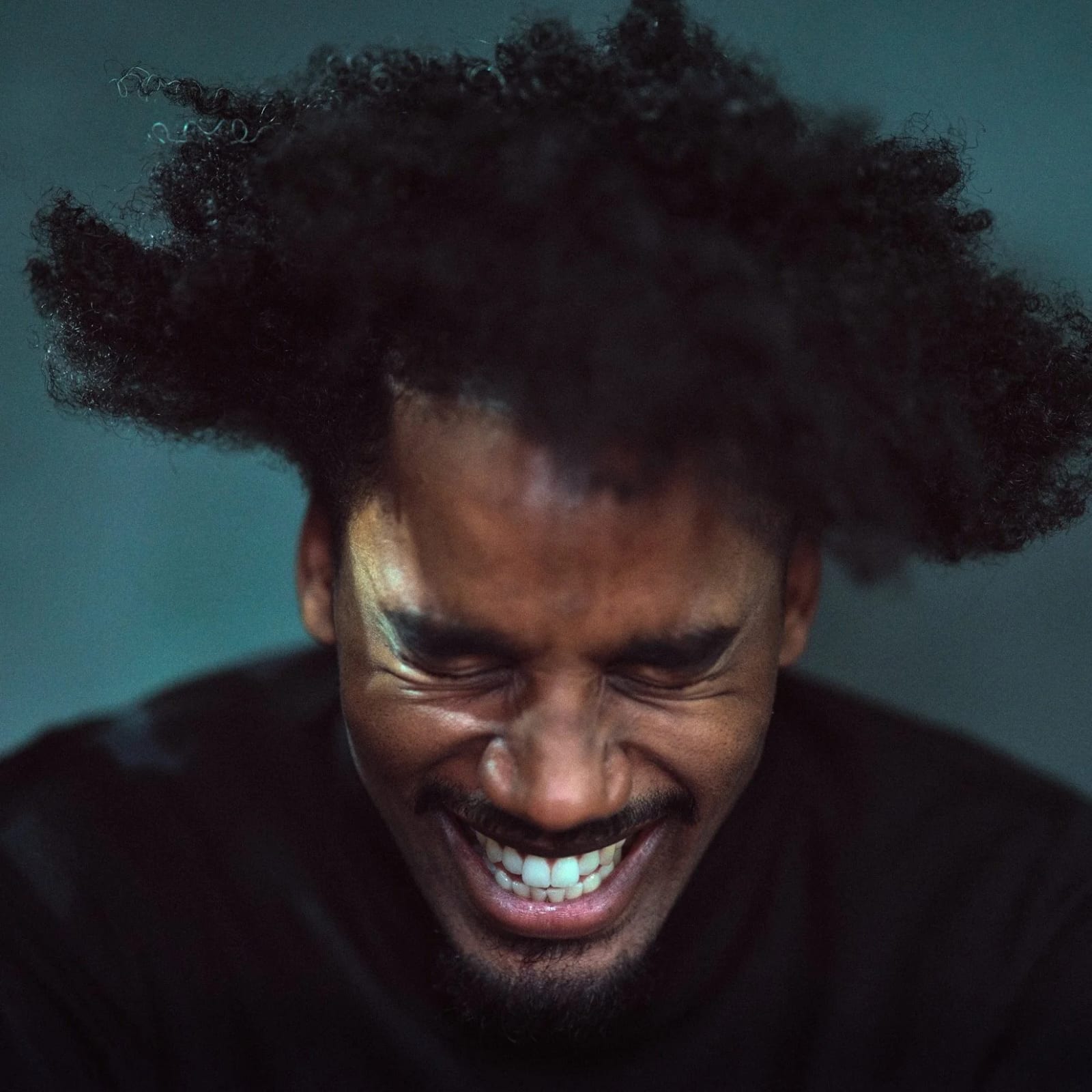 A man laughing with his eyes squeezed shut, his hair flung upward, lit in teal light against a dark background.