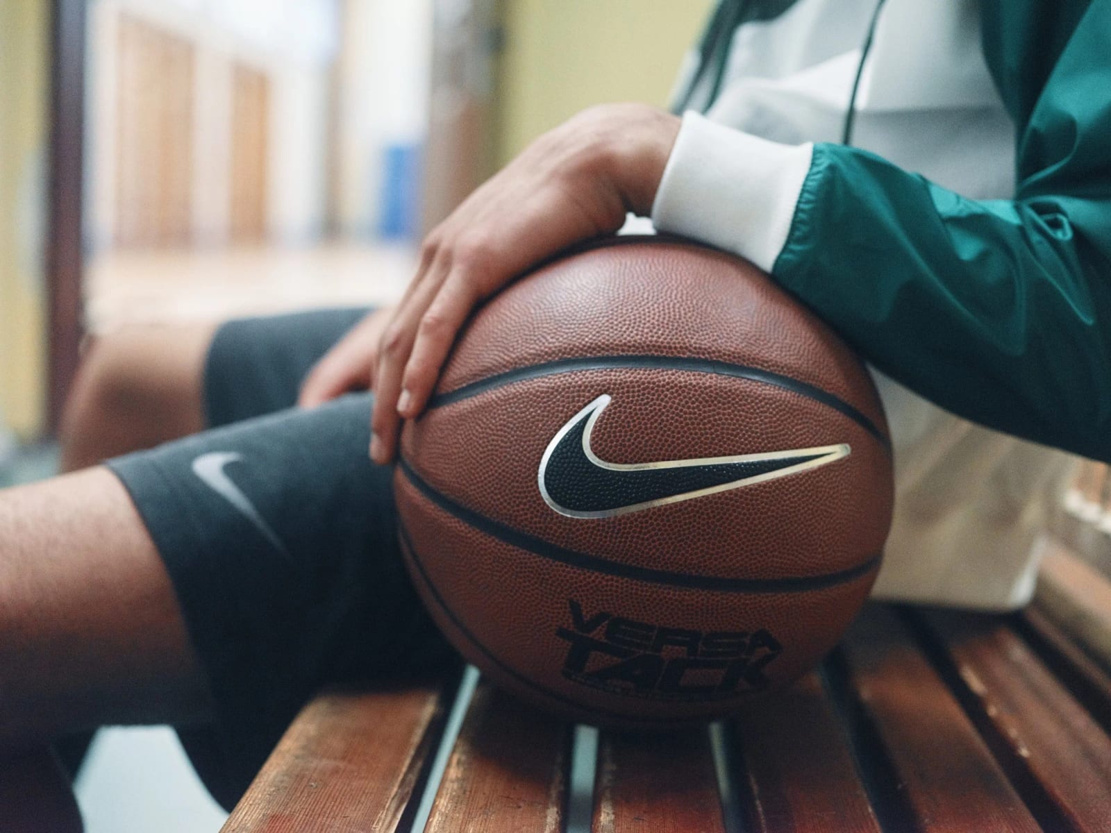 Close-up of a hand resting on a Nike basketball on a wooden bench, the sleeve of a green and white track jacket at the edge of the frame.