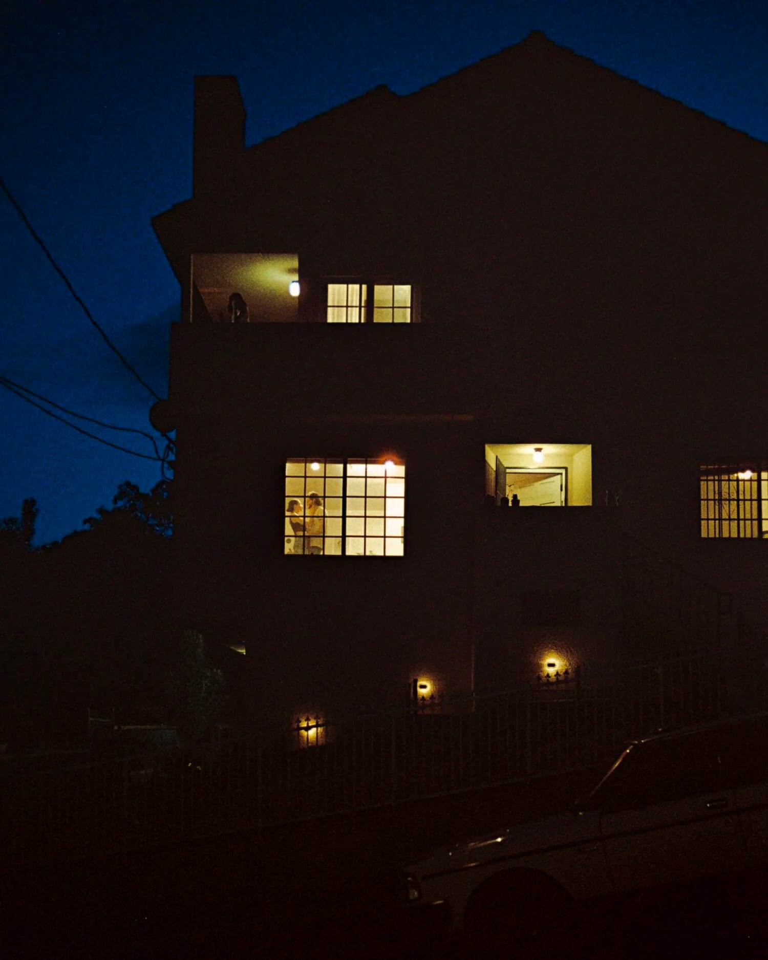A house at night with lit windows, people visible inside, against a deep blue sky.