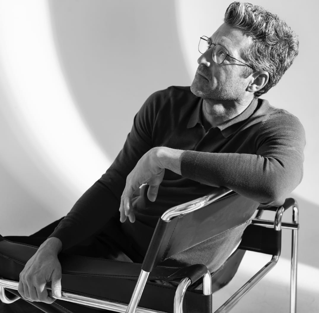 Black and white photograph of a grey-haired man in glasses and a dark sweater sitting in a leather sling chair, looking up.
