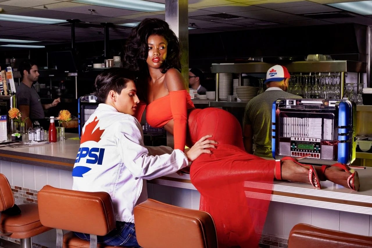 A woman in an orange dress lying across a diner counter while a man in a Pepsi jacket looks on, a jukebox behind.