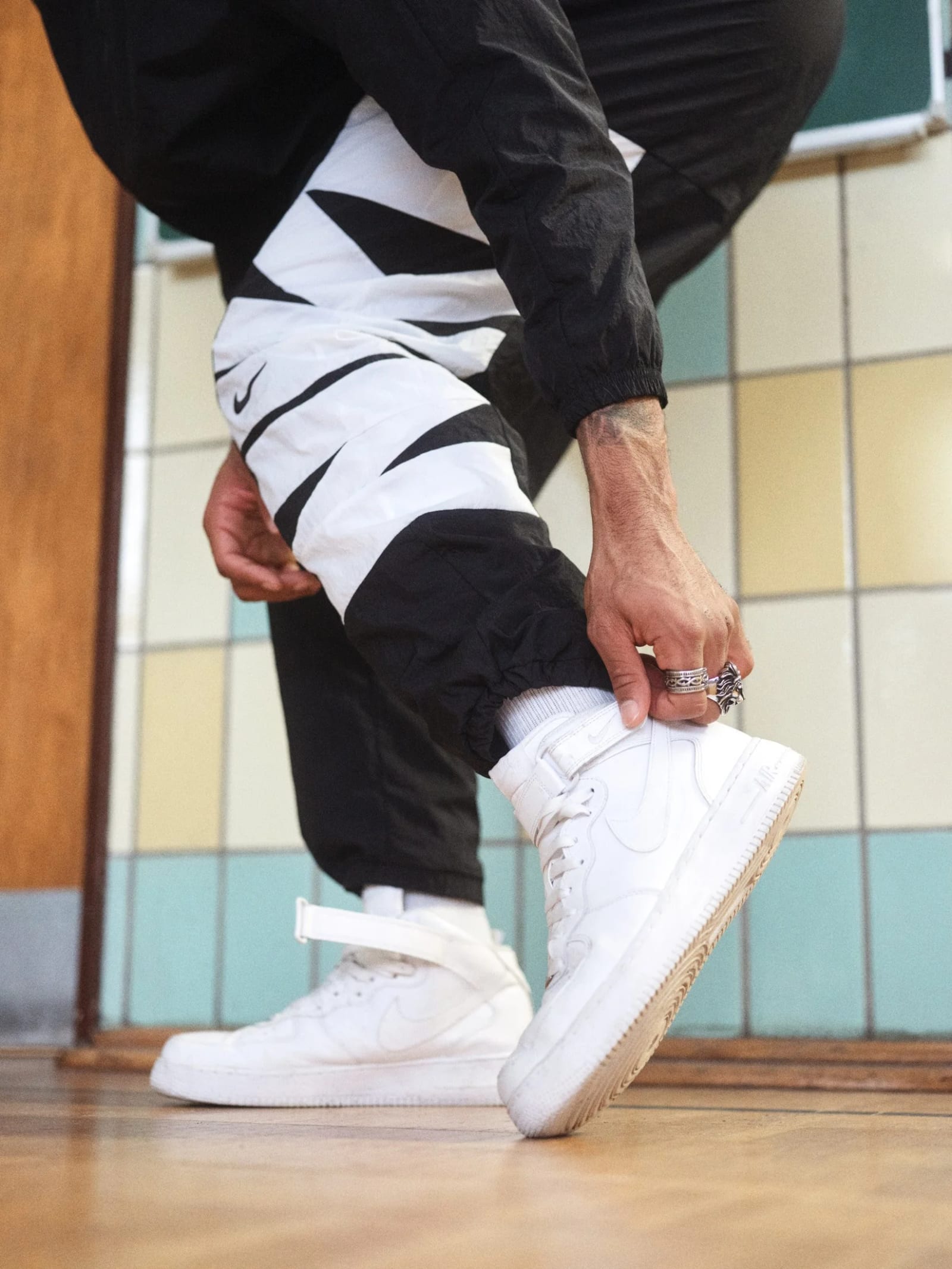 Low angle of a man in black and white patterned trousers tying white high-top trainers on a tiled sports-hall floor.