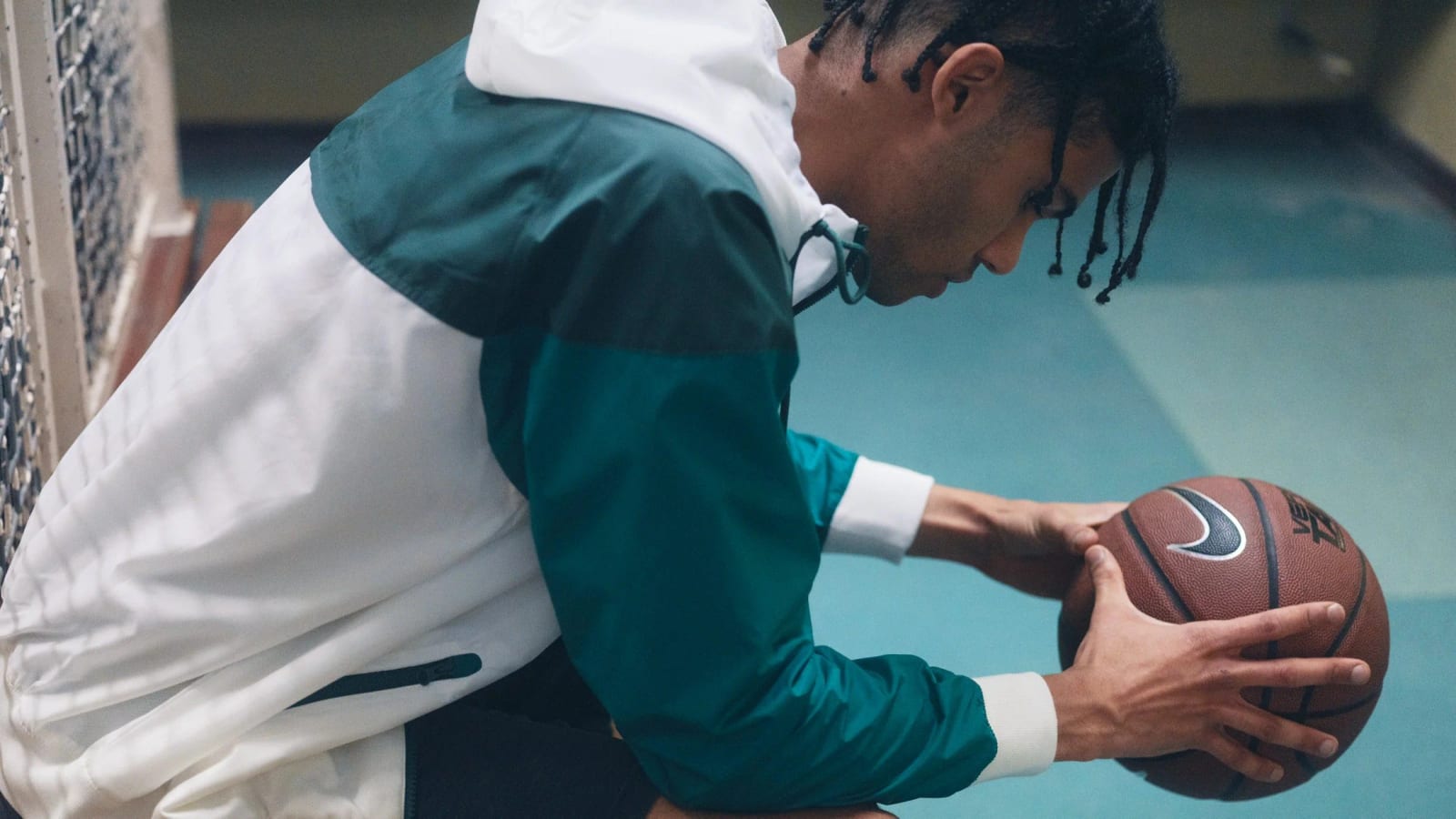 A man in a green and white track jacket bent over a basketball on a court floor.