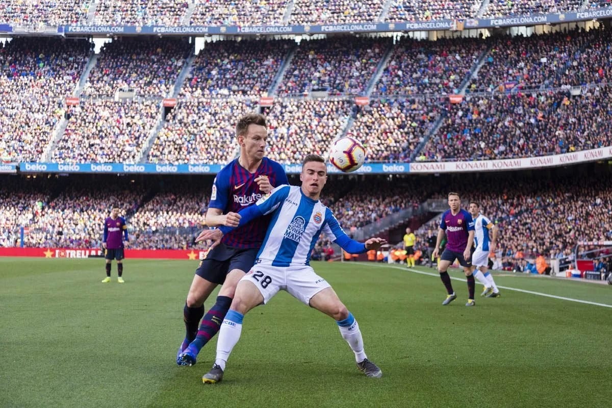 Two footballers, one in blue and red and one in blue and white, contesting a ball in front of a full stadium.