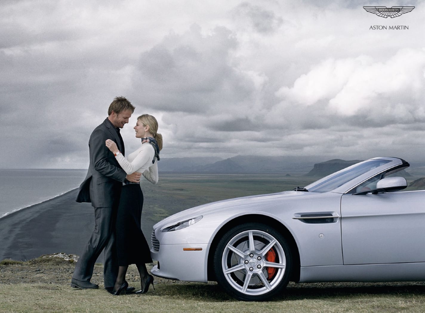 Aston Martin advertisement: a couple embracing beside a silver convertible on a cliff above the sea.