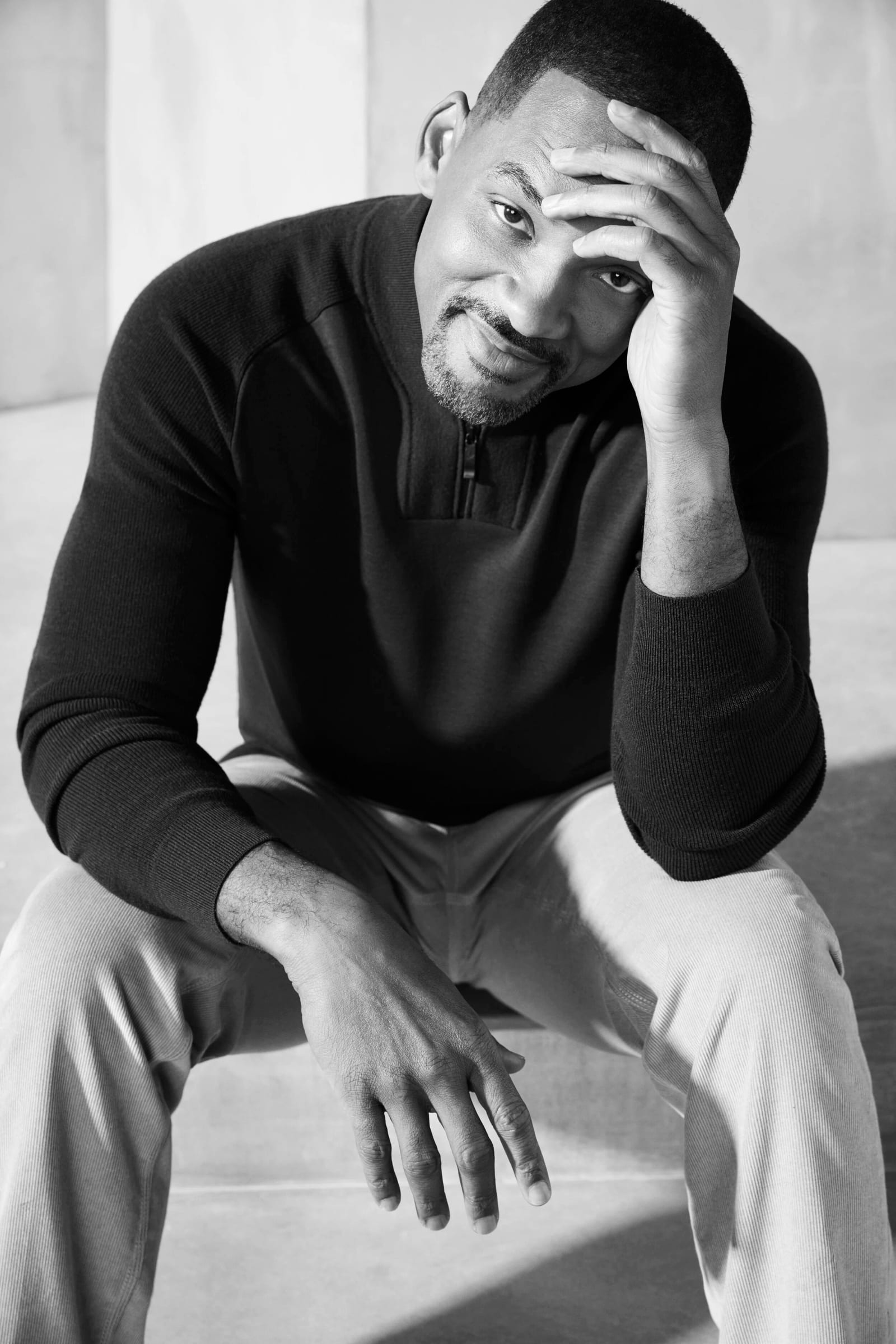 Black and white photograph of Will Smith in a dark zip-neck sweater sitting with one hand on his forehead, smiling.