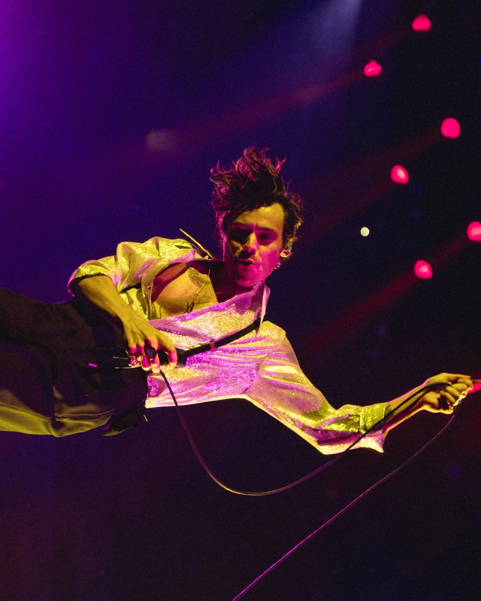 Harry Styles in a metallic suit leaping mid-air on stage under purple and yellow light, microphone in hand.