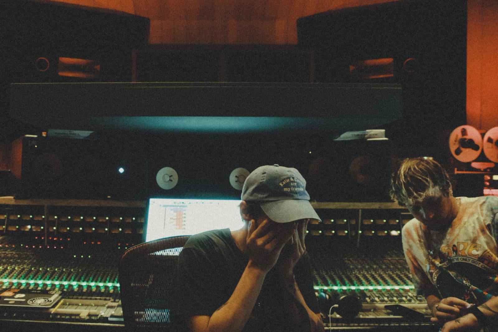 A man in a white cap holding his head at a studio mixing desk, another man beside him, reel-to-reel tape machines behind.