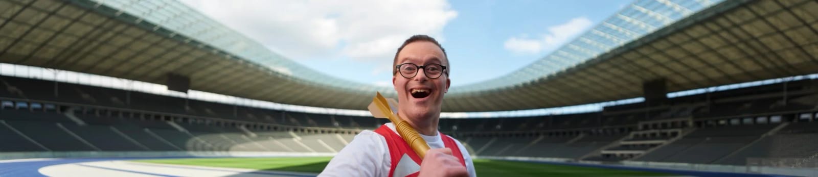 A man in a white and red shirt laughing at the camera in the middle of an empty stadium under a blue sky.