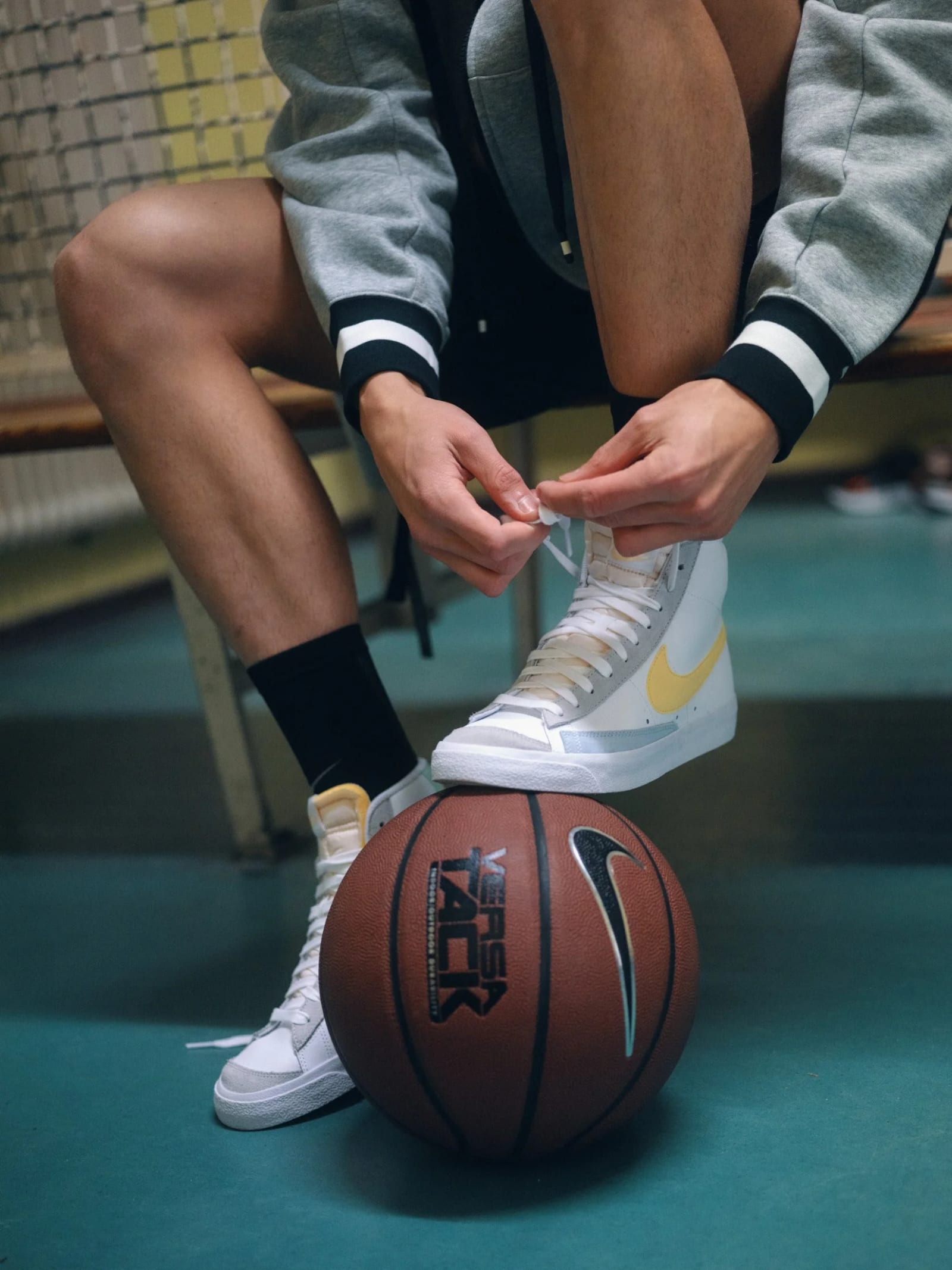 A man in a grey varsity jacket tying white high-top trainers with a yellow swoosh, a basketball under his foot.