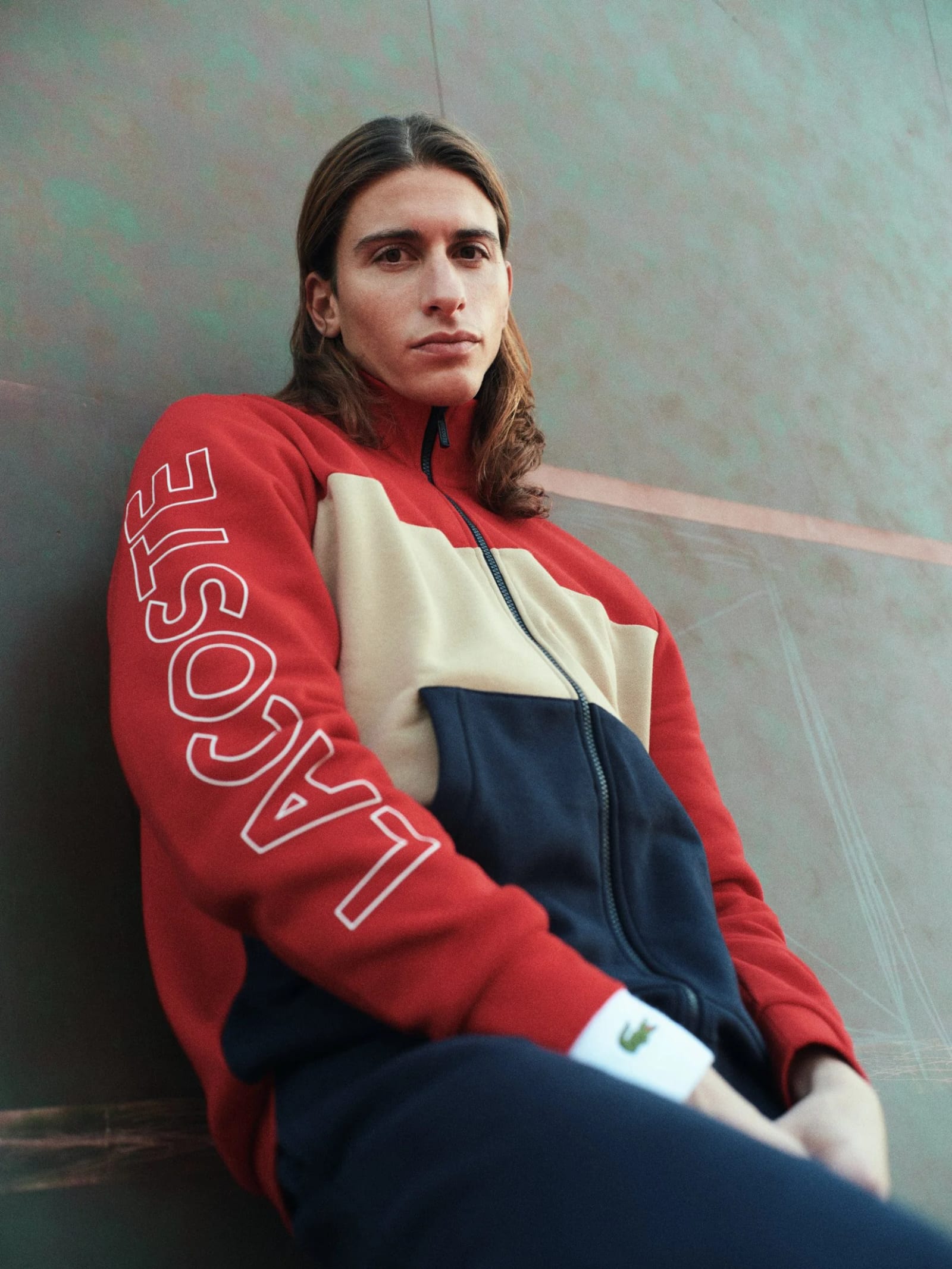 A long-haired man in a red, cream and navy Lacoste track jacket leaning against a green wall.