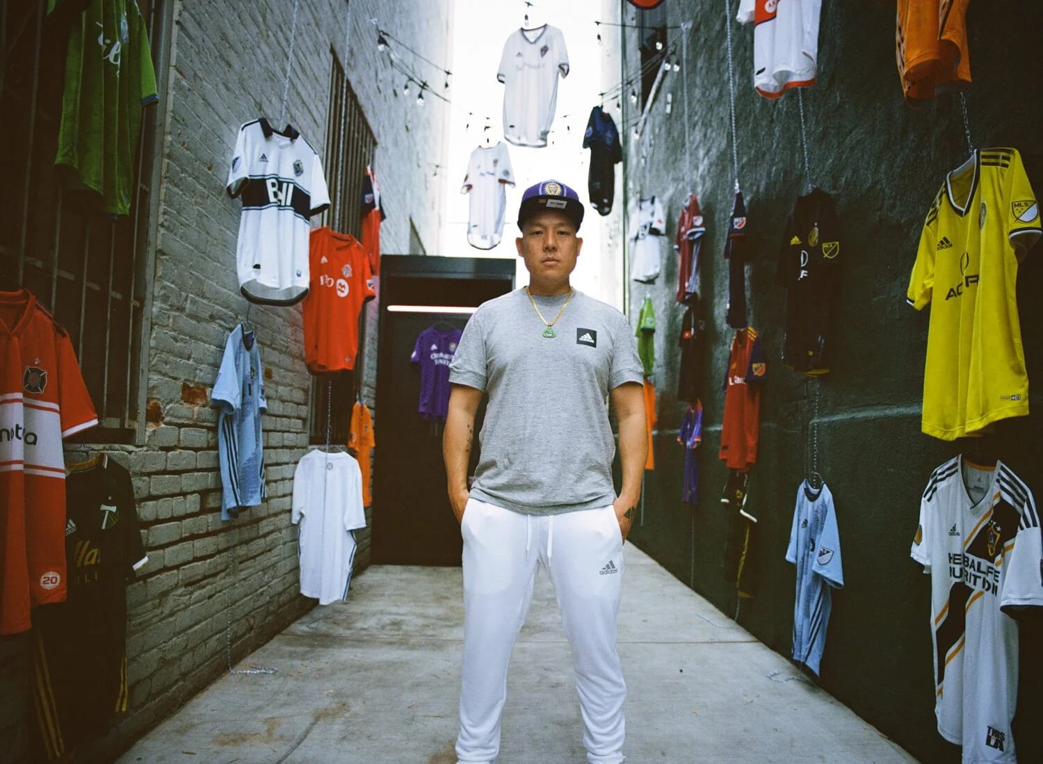 A man in a grey T-shirt and cap standing in an alley hung with football shirts on both walls.