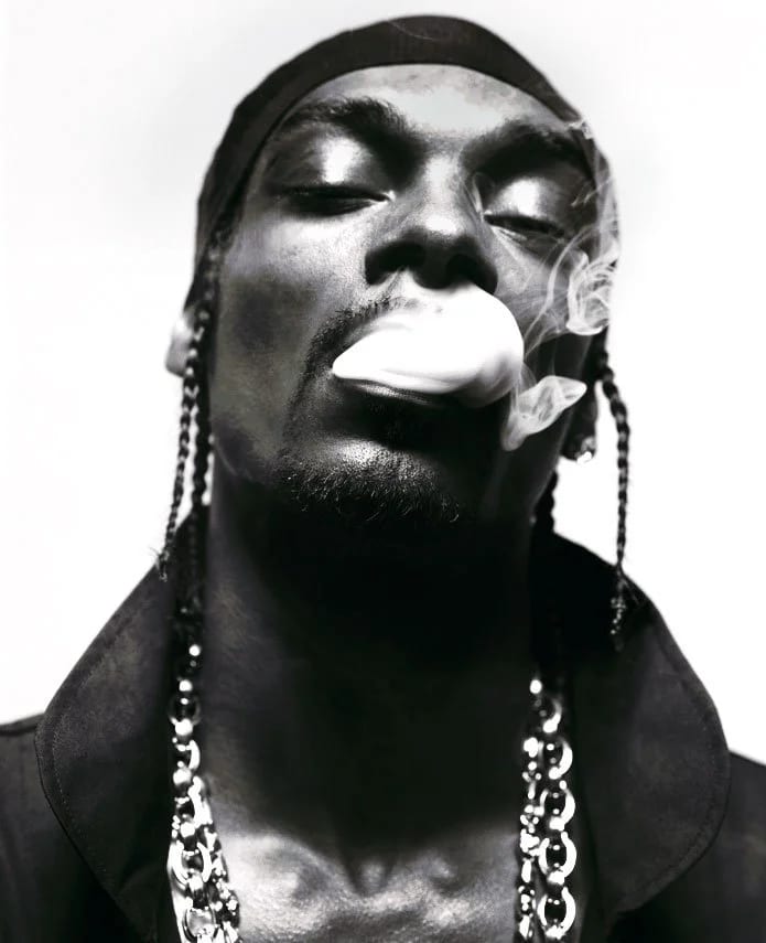 Black and white close-up of a man with braids and a bandana exhaling smoke, a thick chain around his neck.