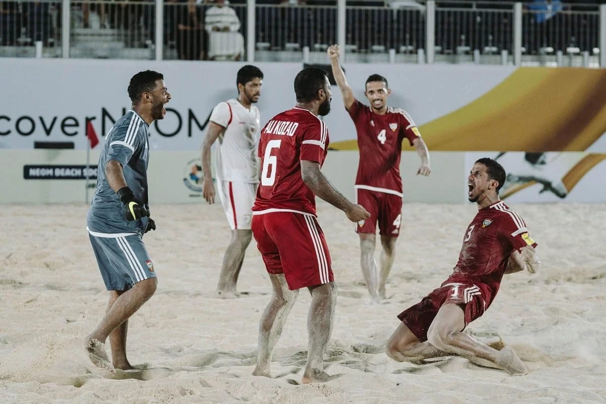 Beach footballers in red and a goalkeeper in blue celebrating on sand, one sliding on his knees, a Neom banner behind.