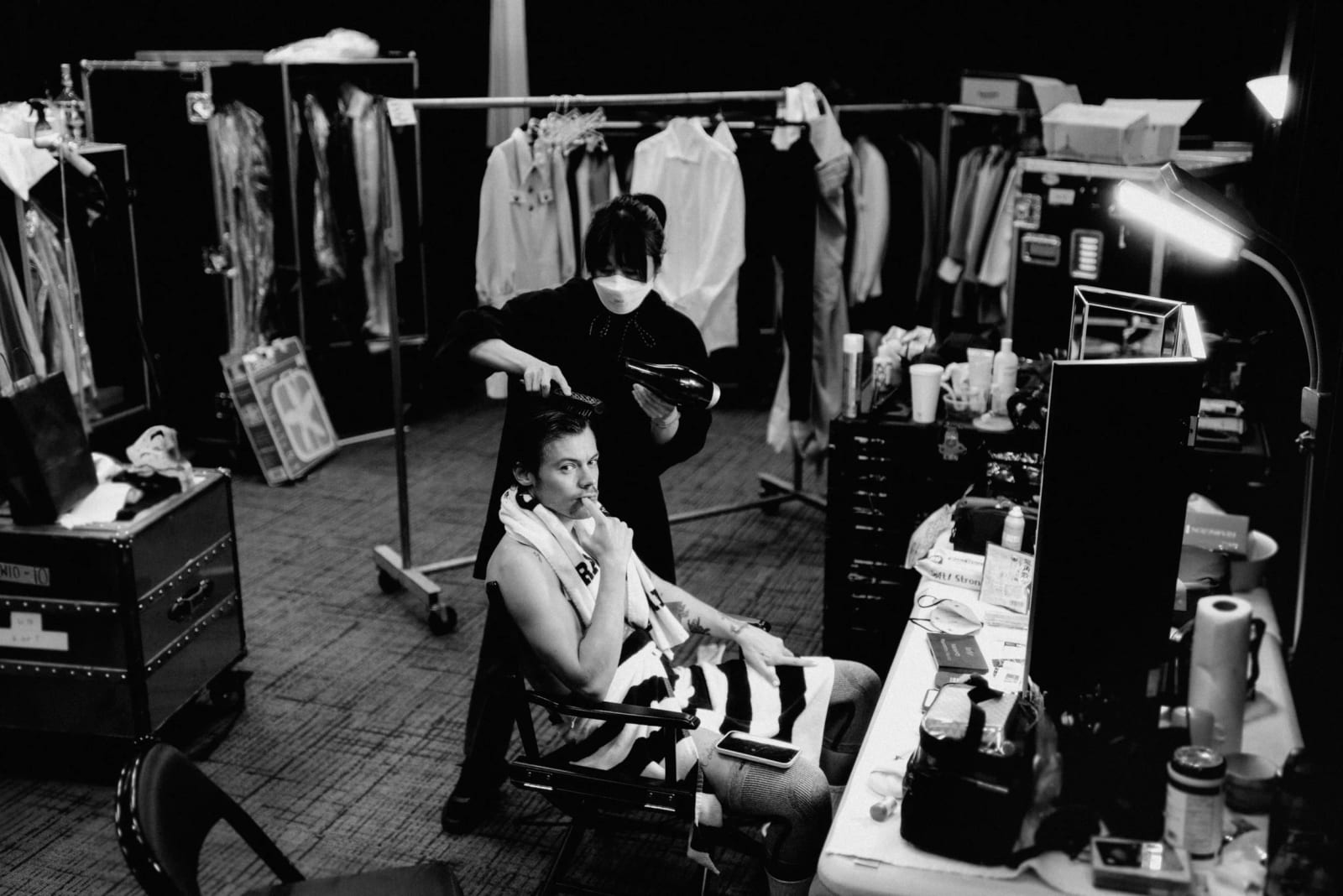 Black and white photograph of a man in a striped top sitting among clothing rails and flight cases while a masked stylist works behind him.