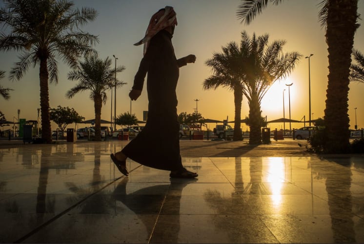 A man in a dark robe and headscarf striding across a wet marble plaza between palm trees at sunset.