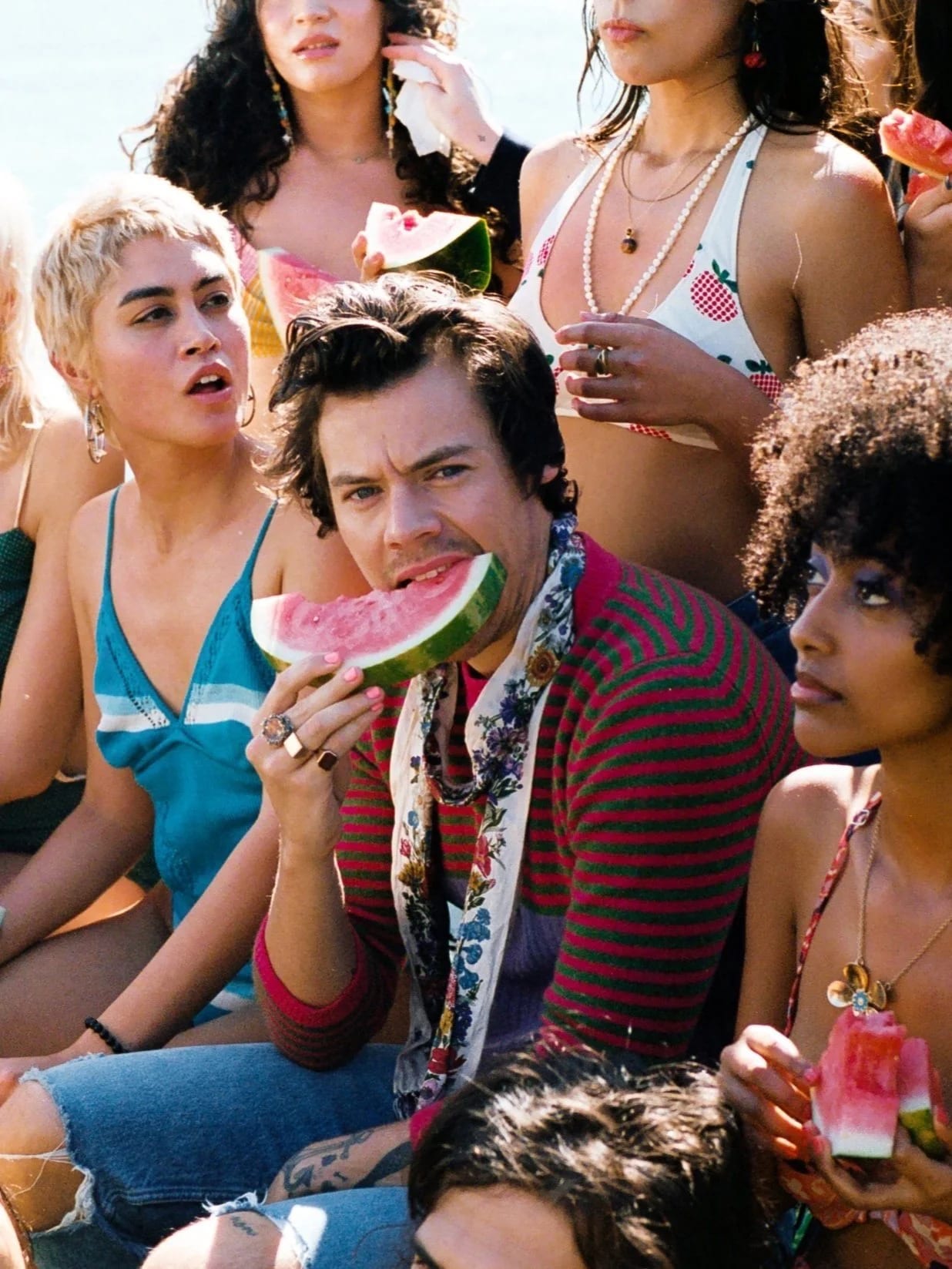 Harry Styles in a striped red cardigan biting into a slice of watermelon, surrounded by women in swimwear.
