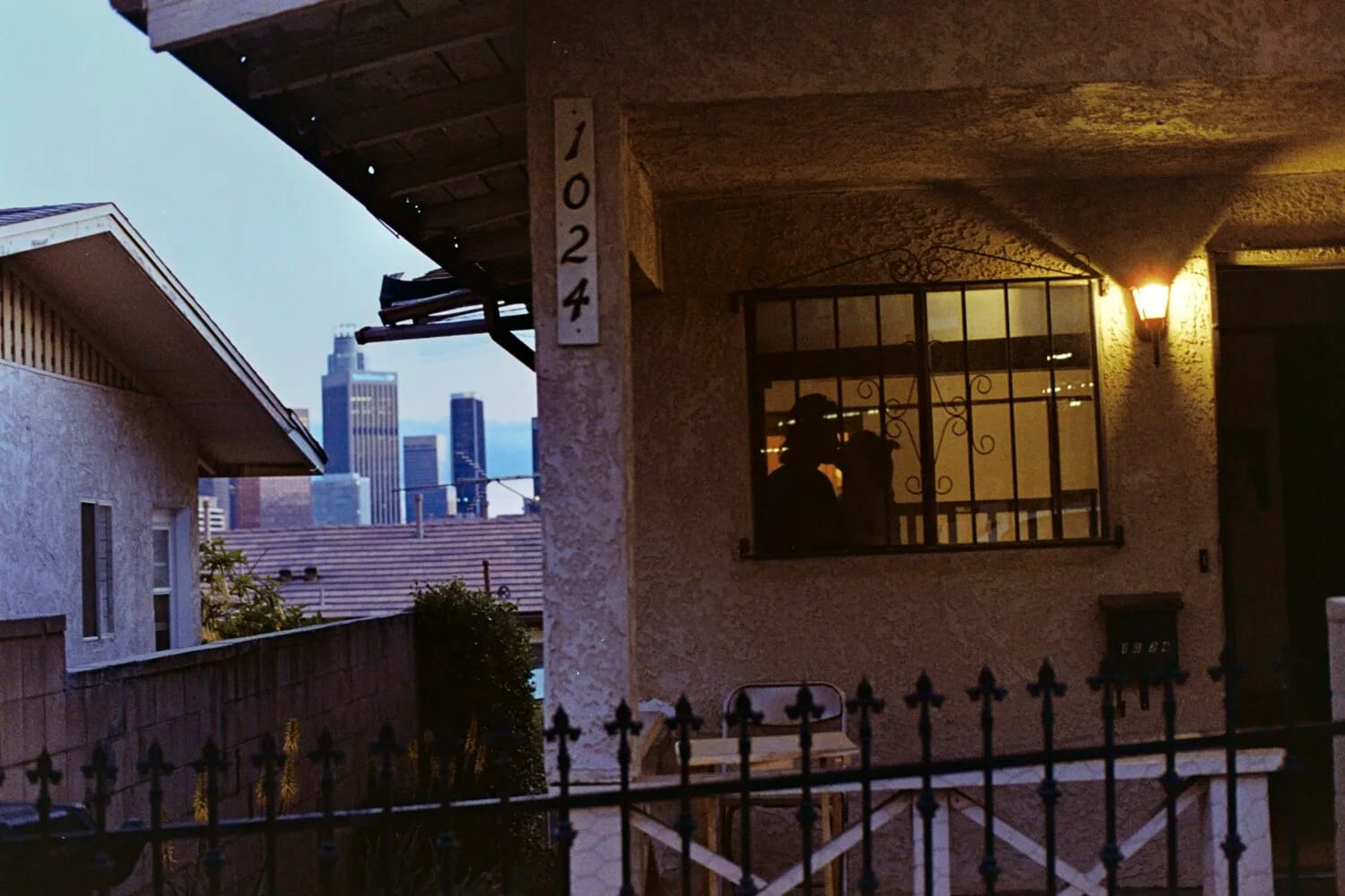 A stucco house numbered 1024 at dusk with a city skyline behind, two figures silhouetted in its barred window.