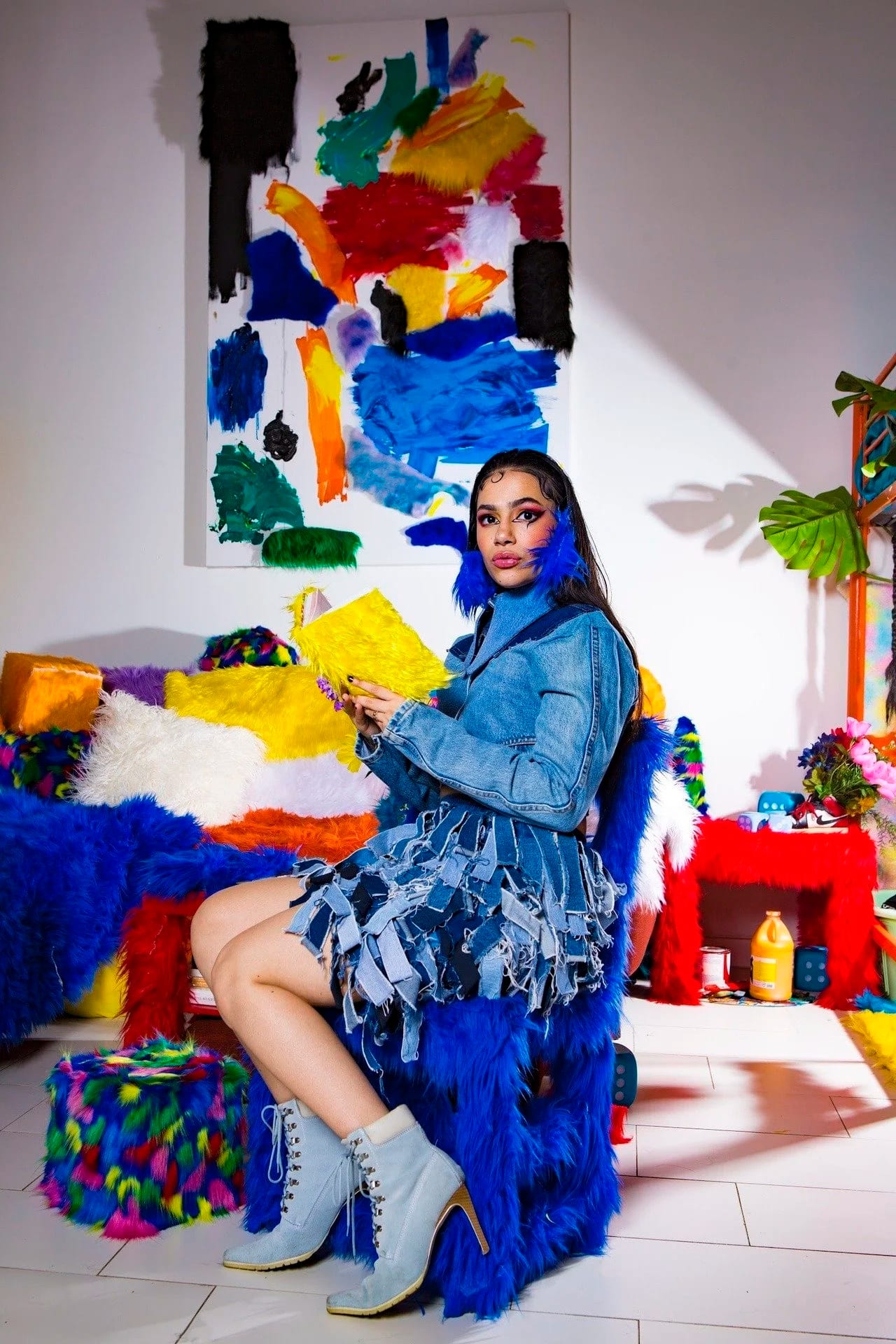 A woman in a denim jacket sitting among furry blue and yellow furniture in a white room with paint-splashed walls.