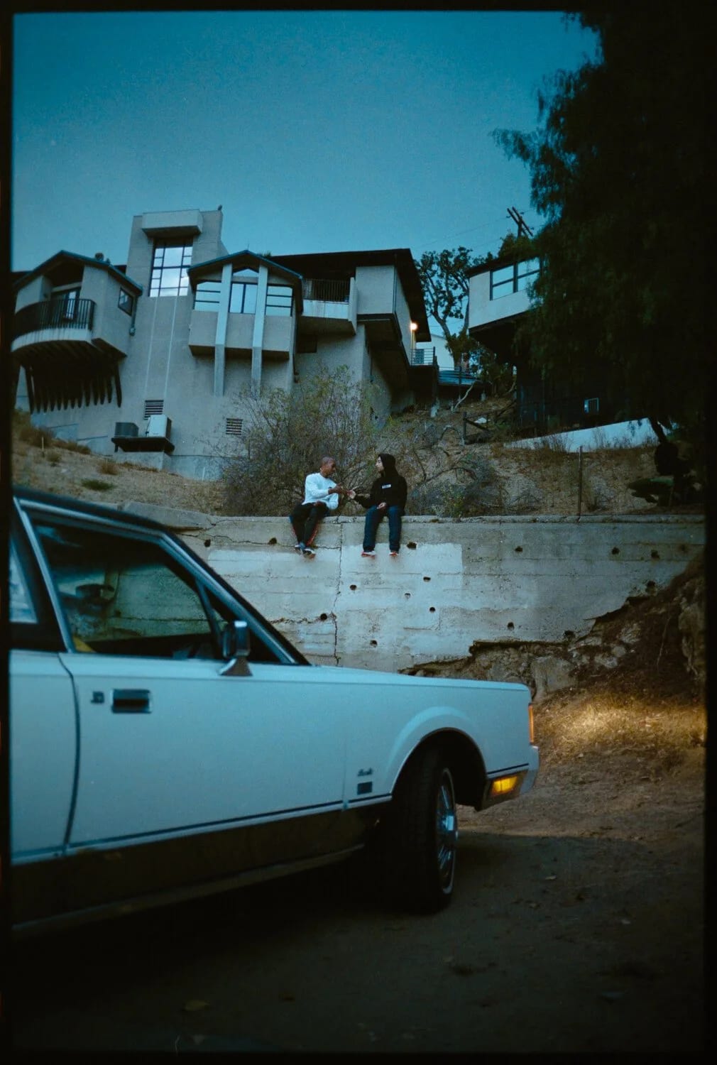 A white car parked below a hillside house at dusk, two people sitting on a wall above it.