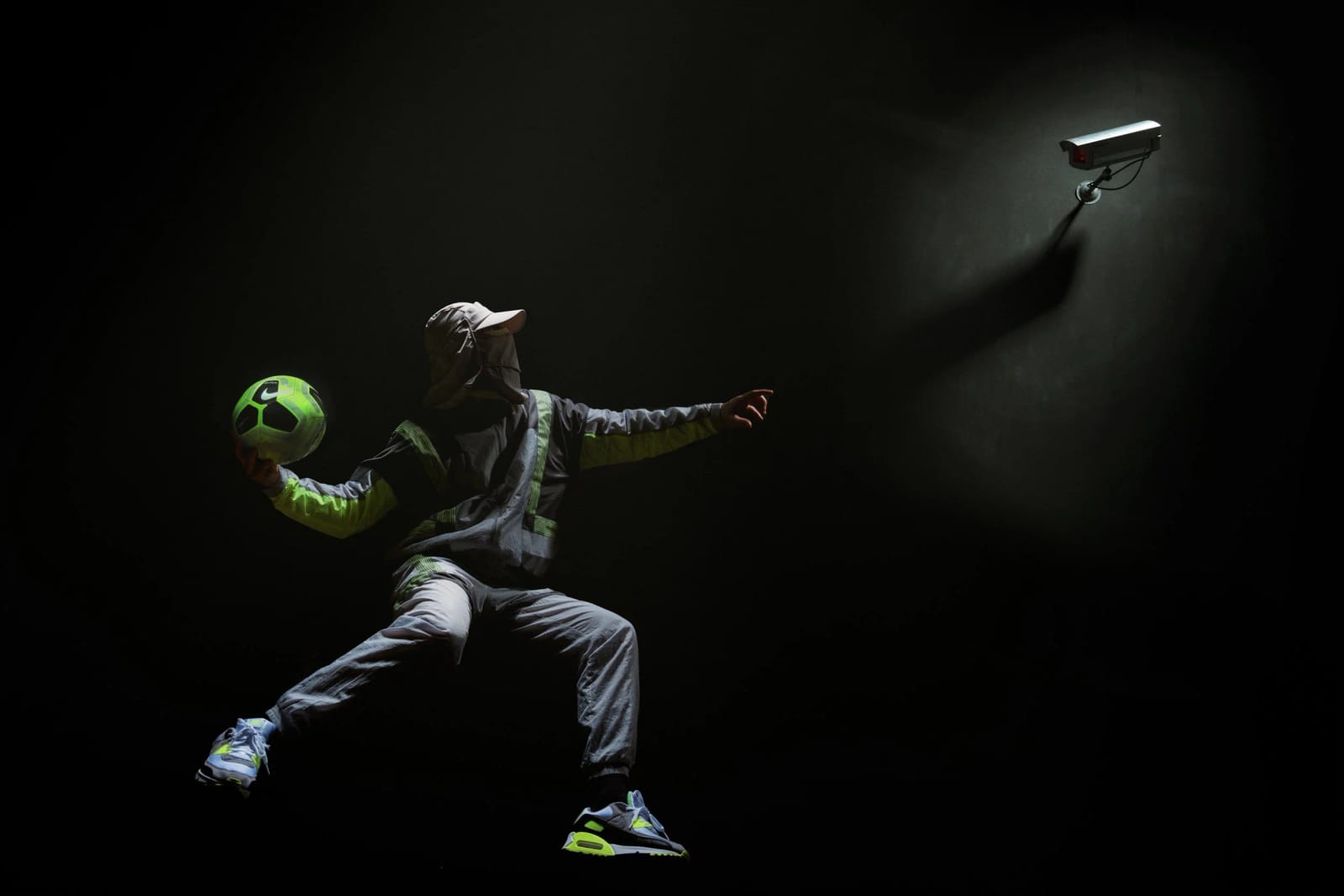 A footballer in a grey tracksuit kicking a bright green ball in a dark space, a security camera lit on the wall behind.