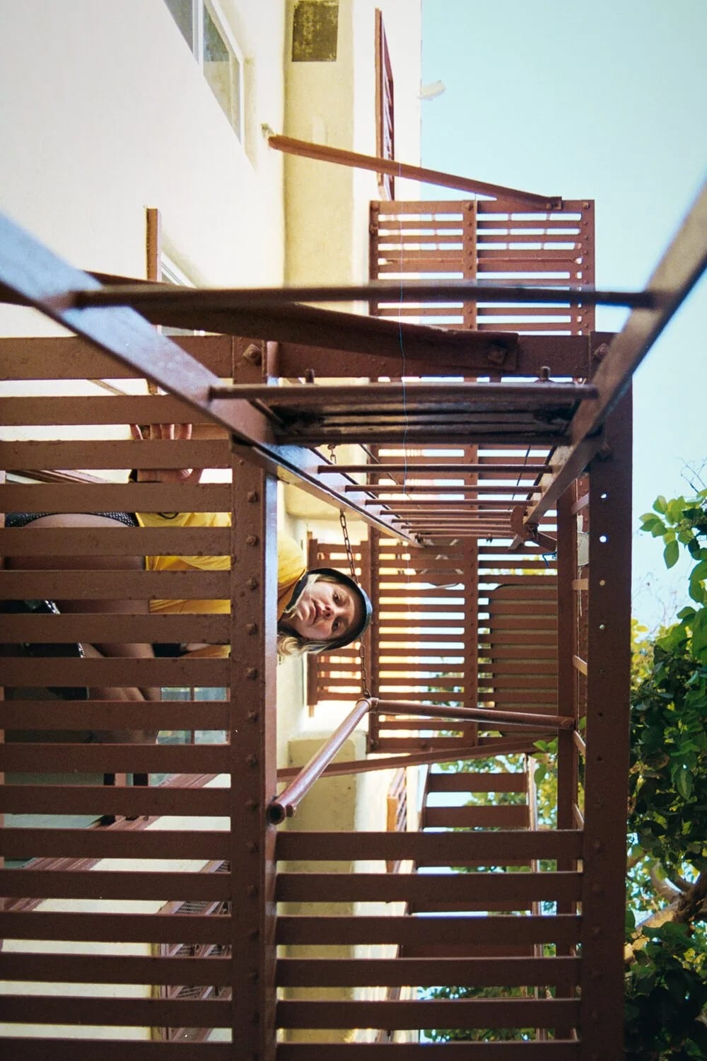 A person peering out from behind the slats of a wooden fire escape on a sunlit building.