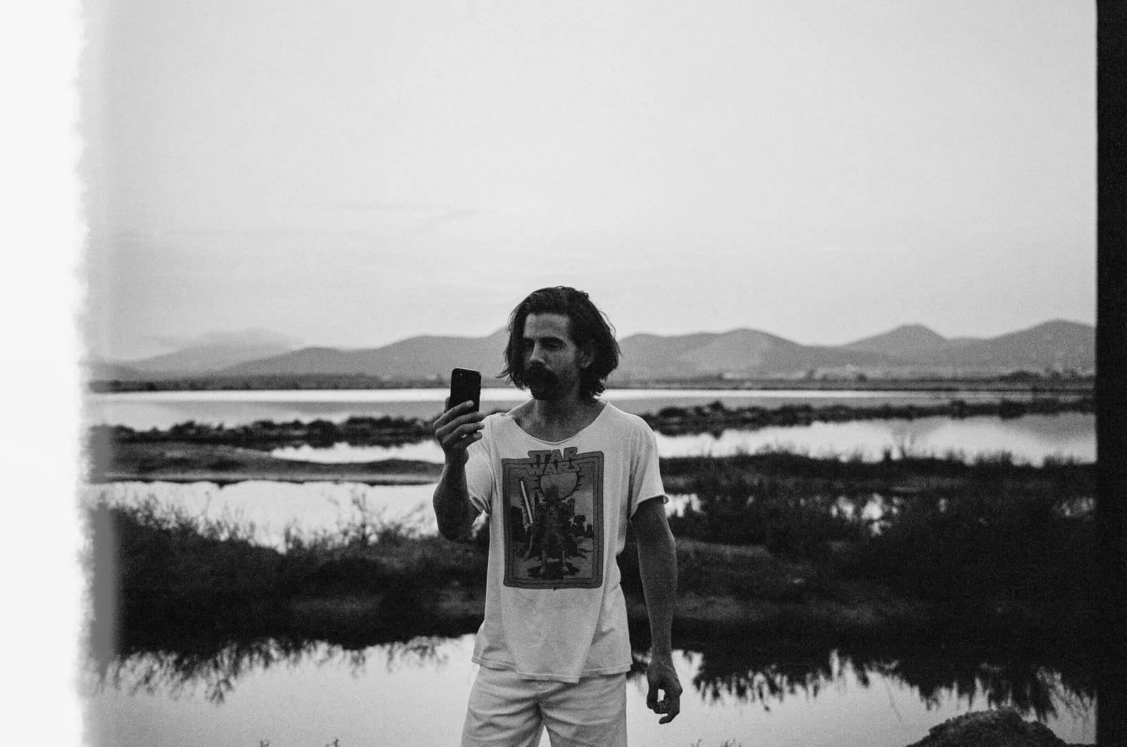 Black and white photograph of Phillip Lopez in a white T-shirt holding up a phone in front of a marsh and mountains.