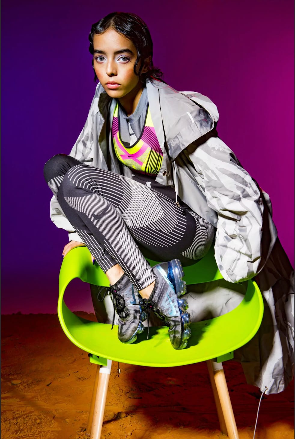 A young woman in grey and neon sportswear crouching on a green chair against purple.