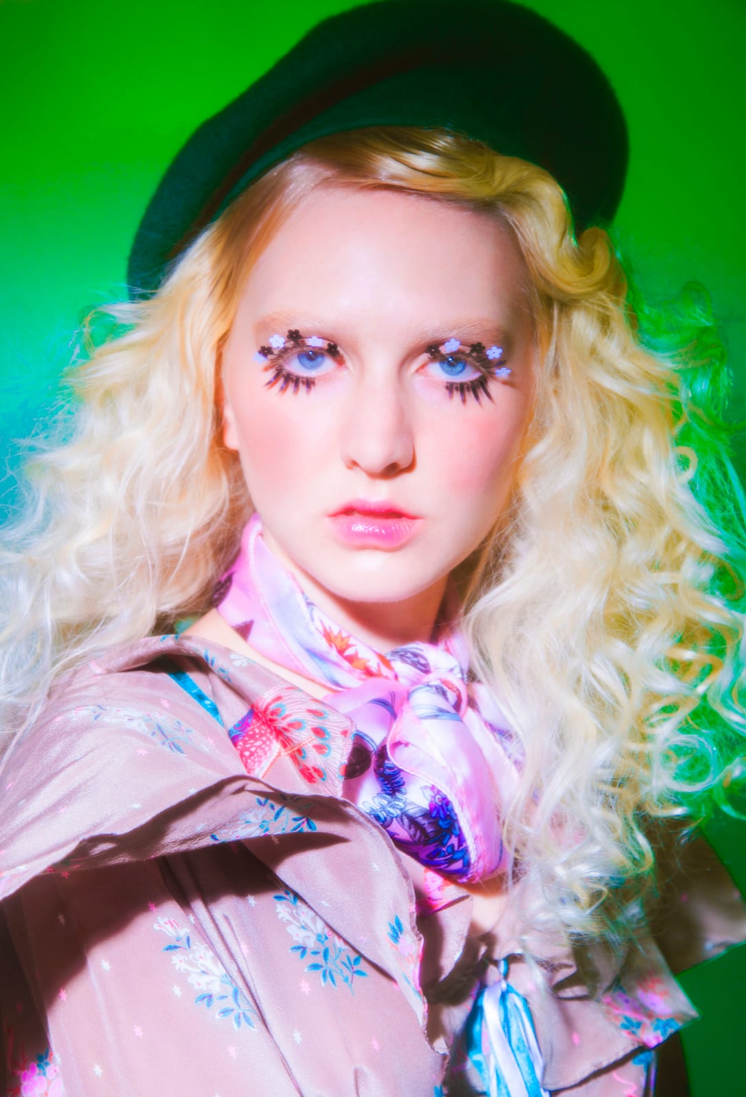 A blonde woman in a green beret and floral blouse with heavy false lashes against green.