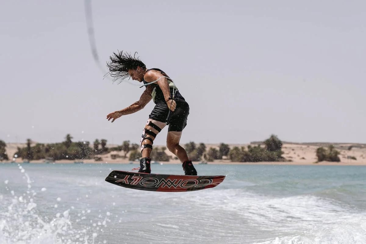 A wakeboarder with long hair carving a spray across flat water, desert dunes and palms behind.