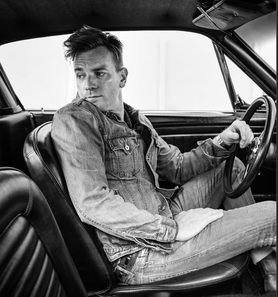 Black and white photograph of Ewan McGregor in a denim jacket sitting at the wheel of a classic car, looking out.