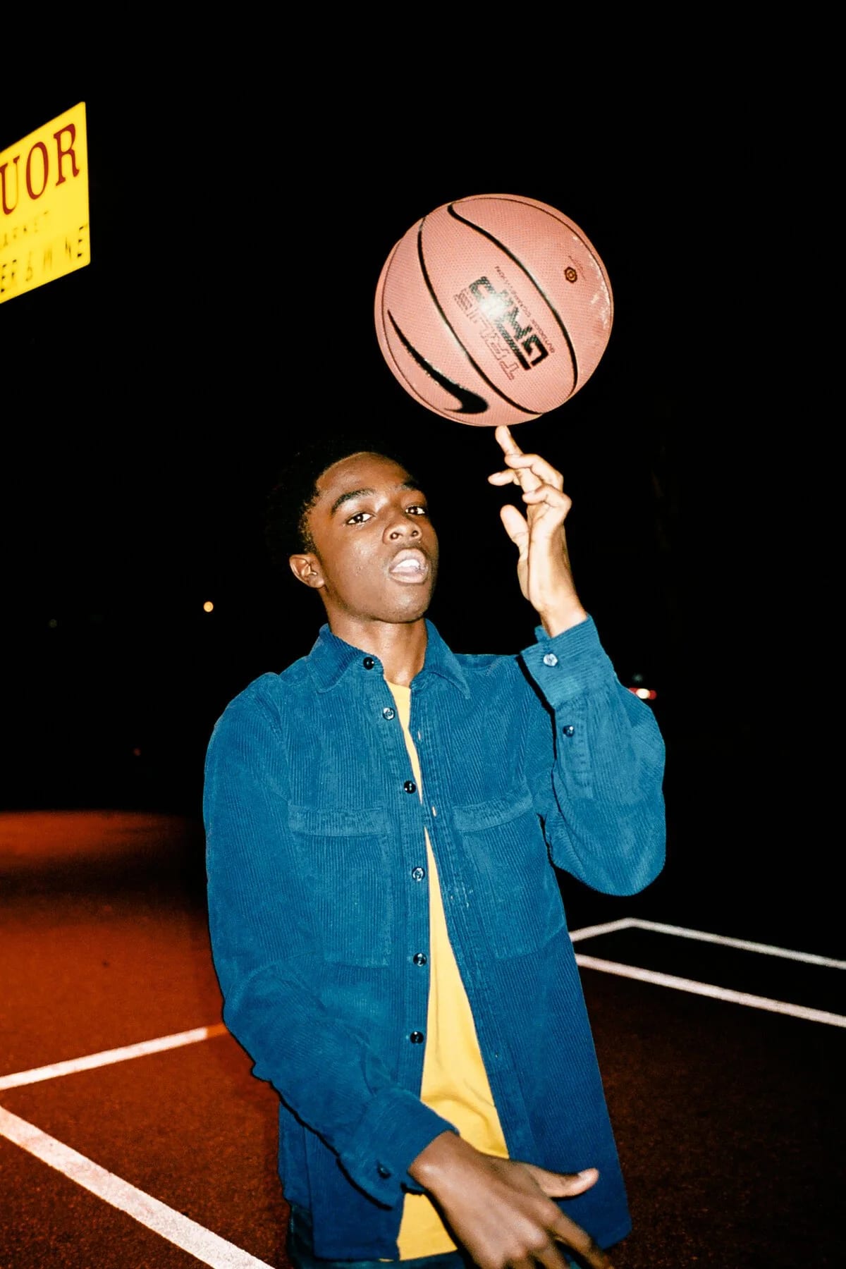A young man in a blue shirt spinning a basketball on his finger at night, a Liquor sign above him.