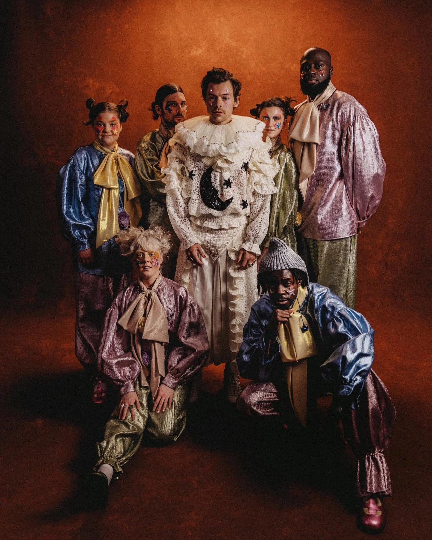 Harry Styles in a white ruffled clown costume with a moon and stars motif, surrounded by seven performers in satin costumes against a rust backdrop.
