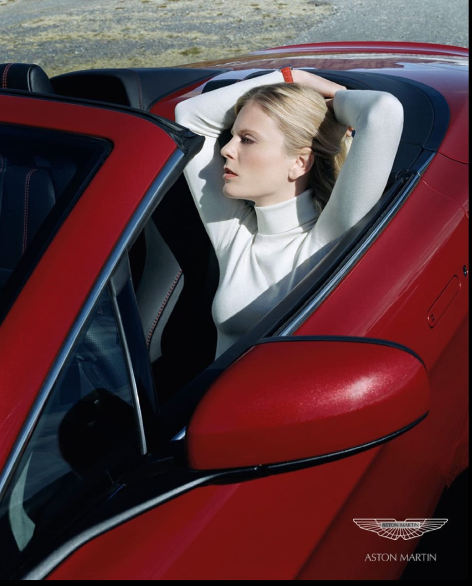 Aston Martin advertisement: a woman in a white turtleneck reclining in the seat of a red convertible with her arms above her head.