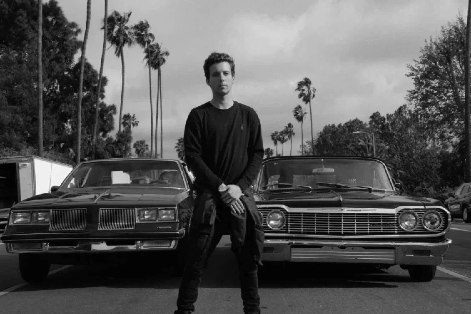 Black and white photograph of Daniel Russell leaning against a vintage car between two others on a palm-lined street.
