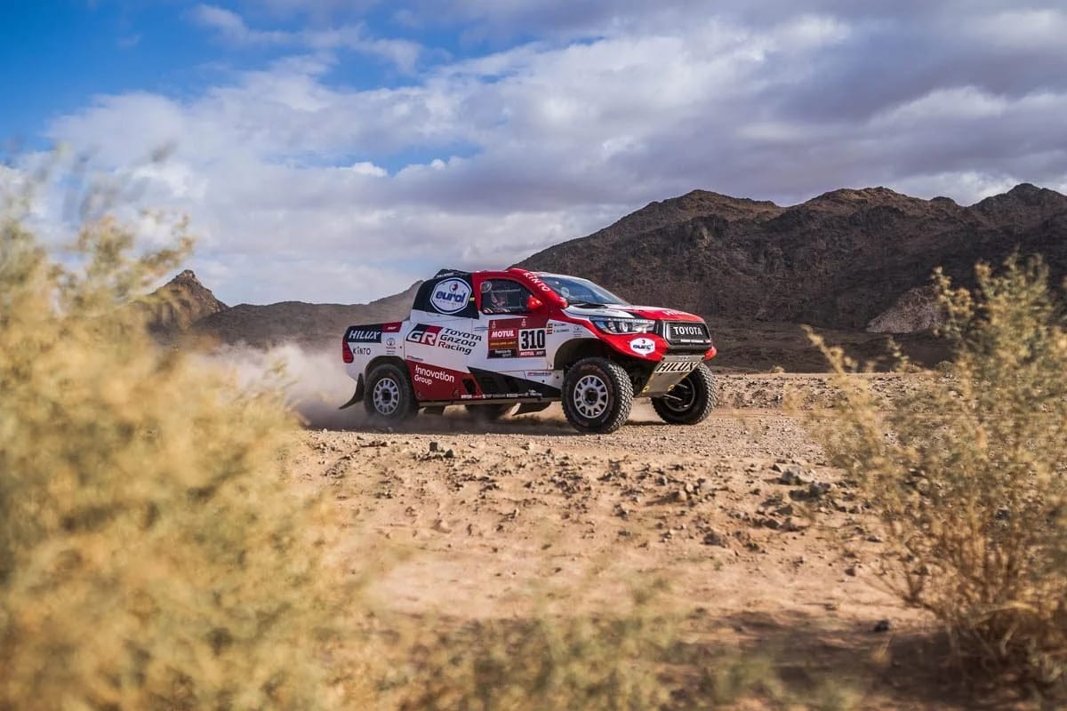 A red and white rally pick-up kicking up dust on a desert track between scrub and mountains.