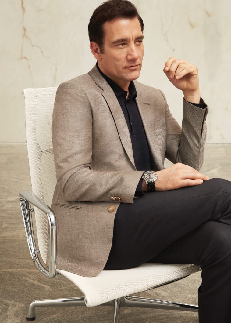 A man in a beige blazer and black shirt sitting in a white chair with his legs crossed.