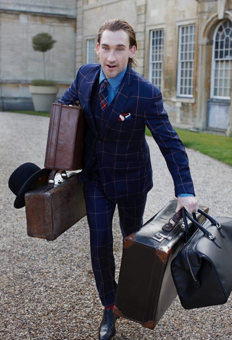 A man in a blue checked suit carrying four vintage suitcases across a gravel drive in front of a stone house.