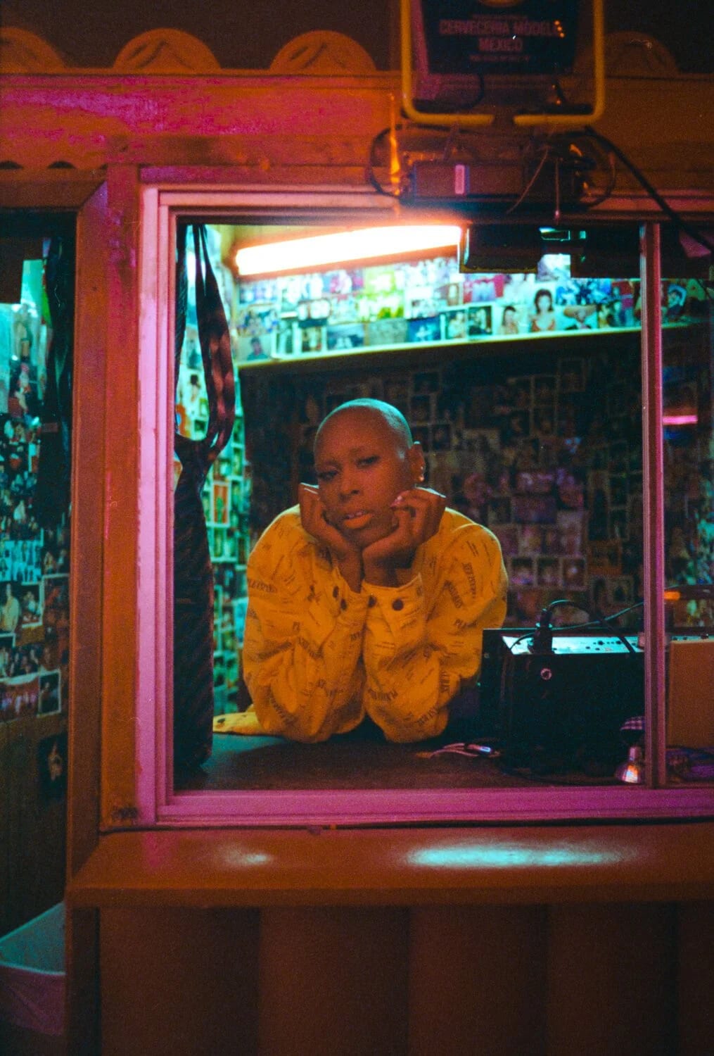 A woman in a yellow sweater resting her chin on her hands at a photo-covered bar window, lit pink.