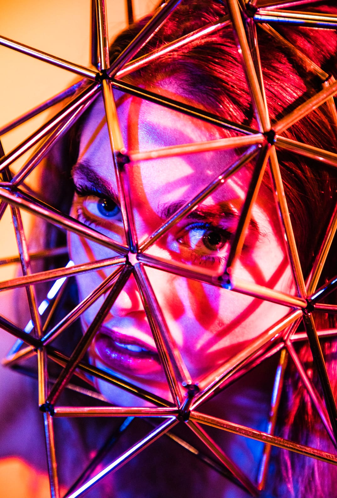 A woman's face seen through a geometric lattice of metal rods lit pink and blue.