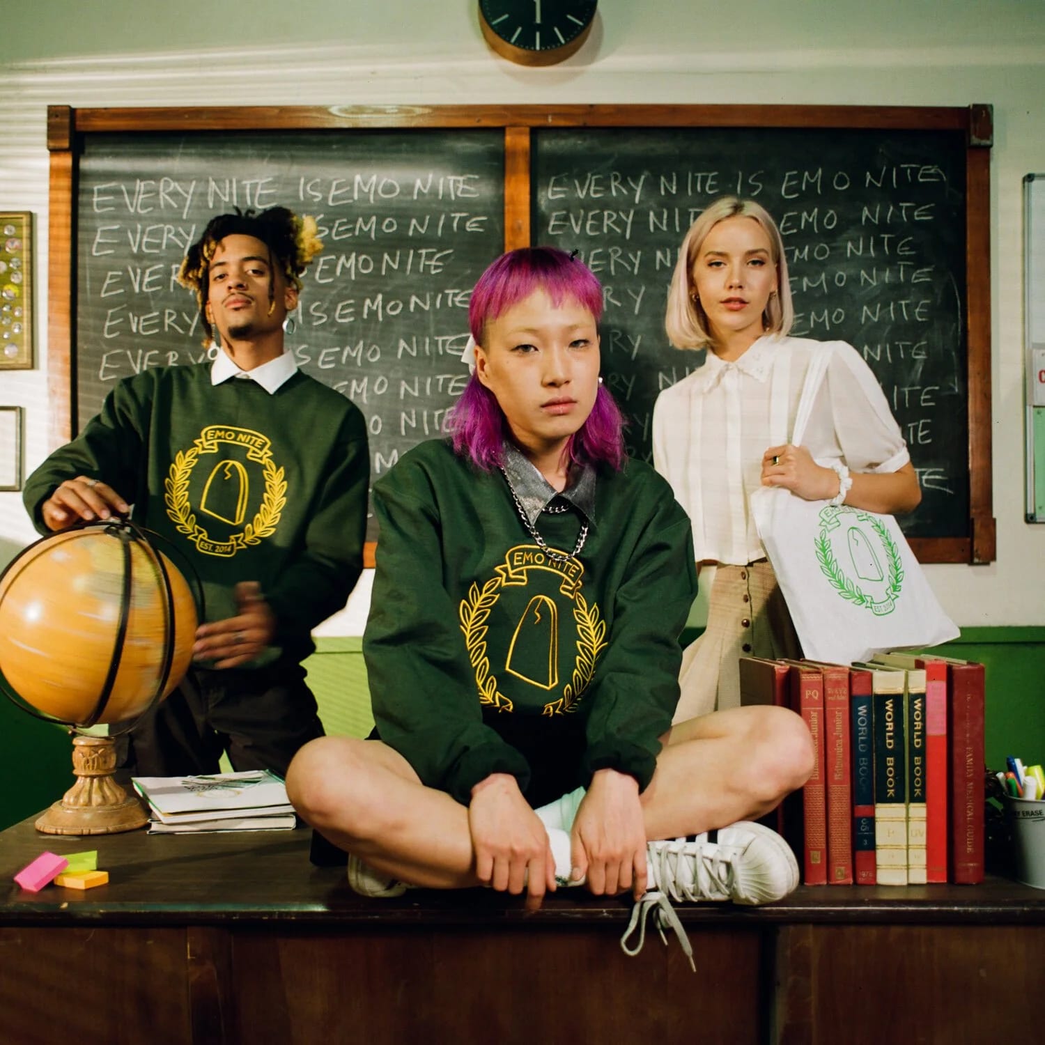 Three people in green sweatshirts and a white shirt sitting on a desk in front of a chalkboard covered in the words Every nite is emo nite, with a globe and a stack of books.