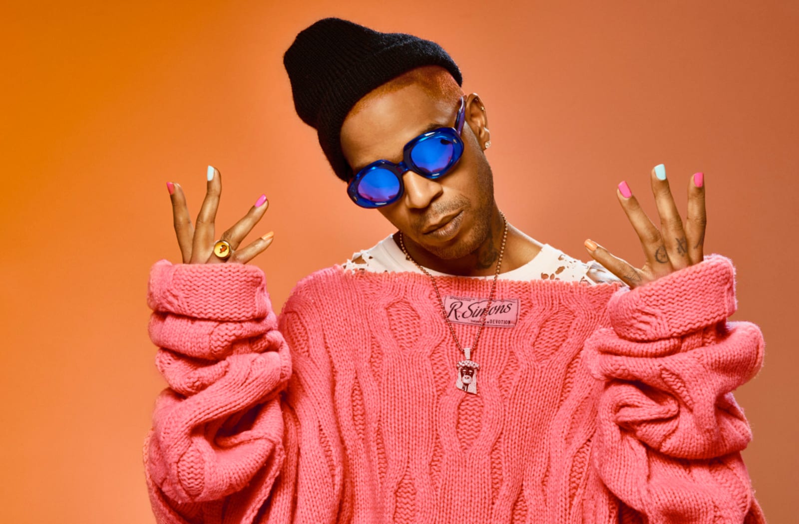 A man in a black beanie, blue sunglasses and a pink knitted sweater holding up his hands with coloured nails, against orange.