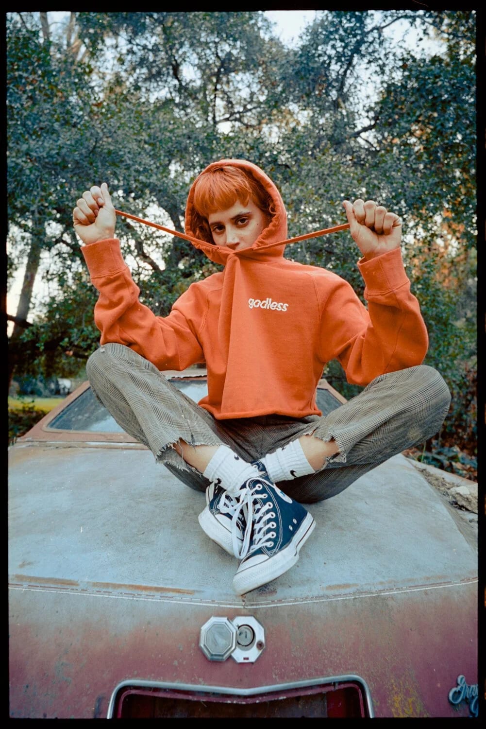 A red-haired young man in an orange hoodie pulling its drawstrings, sitting cross-legged on the bonnet of an old car among trees.
