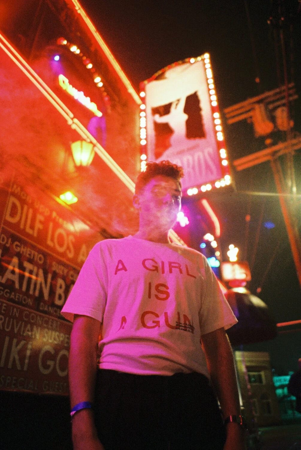 A young man in a white T-shirt reading A girl is a gun standing under neon signs at night, smoke drifting from his mouth.