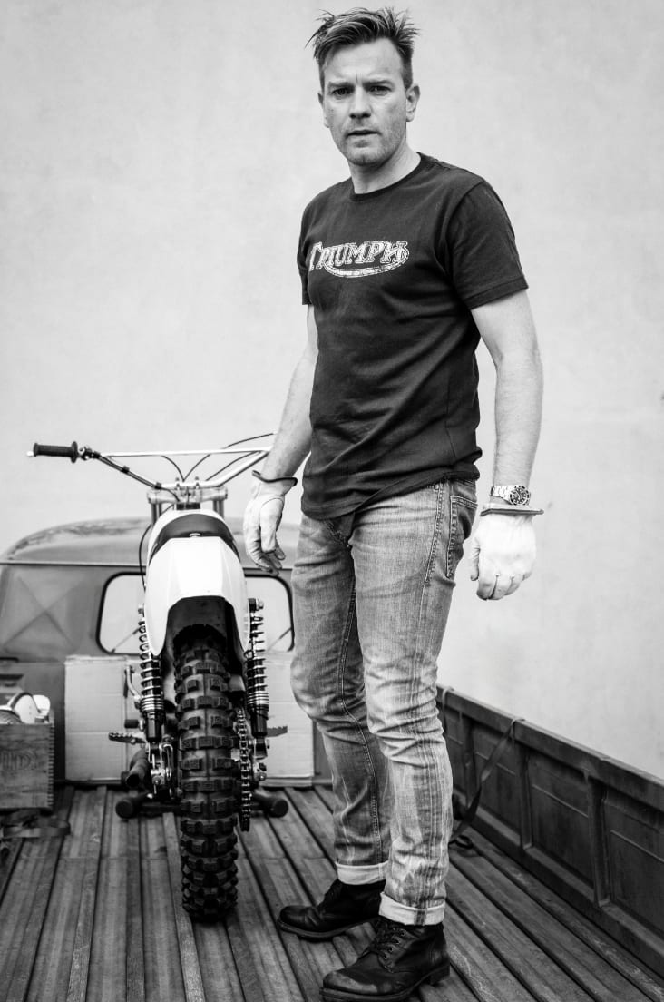 Black and white photograph of Ewan McGregor in a Triumph T-shirt and jeans standing beside a dirt bike.