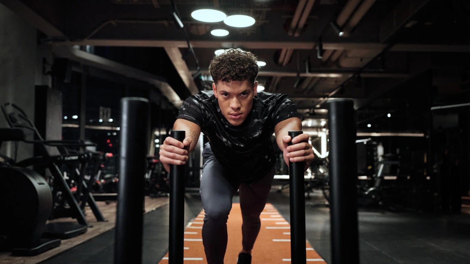 A man in a black top leaning into a weighted sled on a gym track, staring ahead.