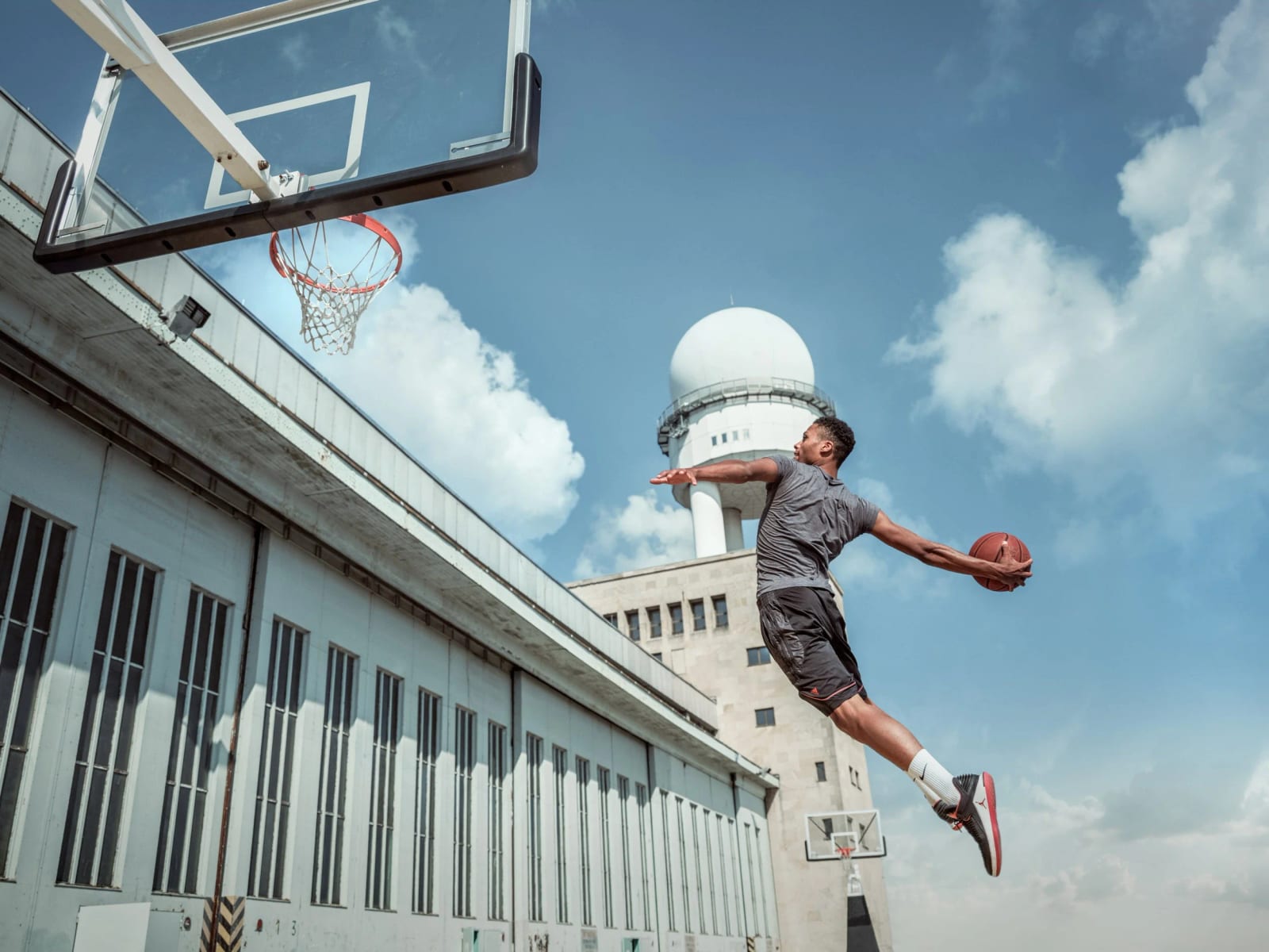 A basketball player leaping toward a hoop in front of a long white hangar building and a domed radar tower under a blue sky.
