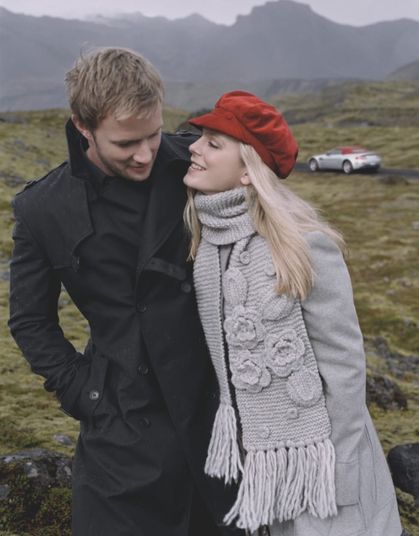 A couple in winter clothes on a moorland road, the woman in a red beret and chunky scarf, a silver sports car behind.