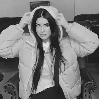 Black and white portrait of Chandler Lass pulling up the hood of a white puffer jacket, sitting in a leather armchair.