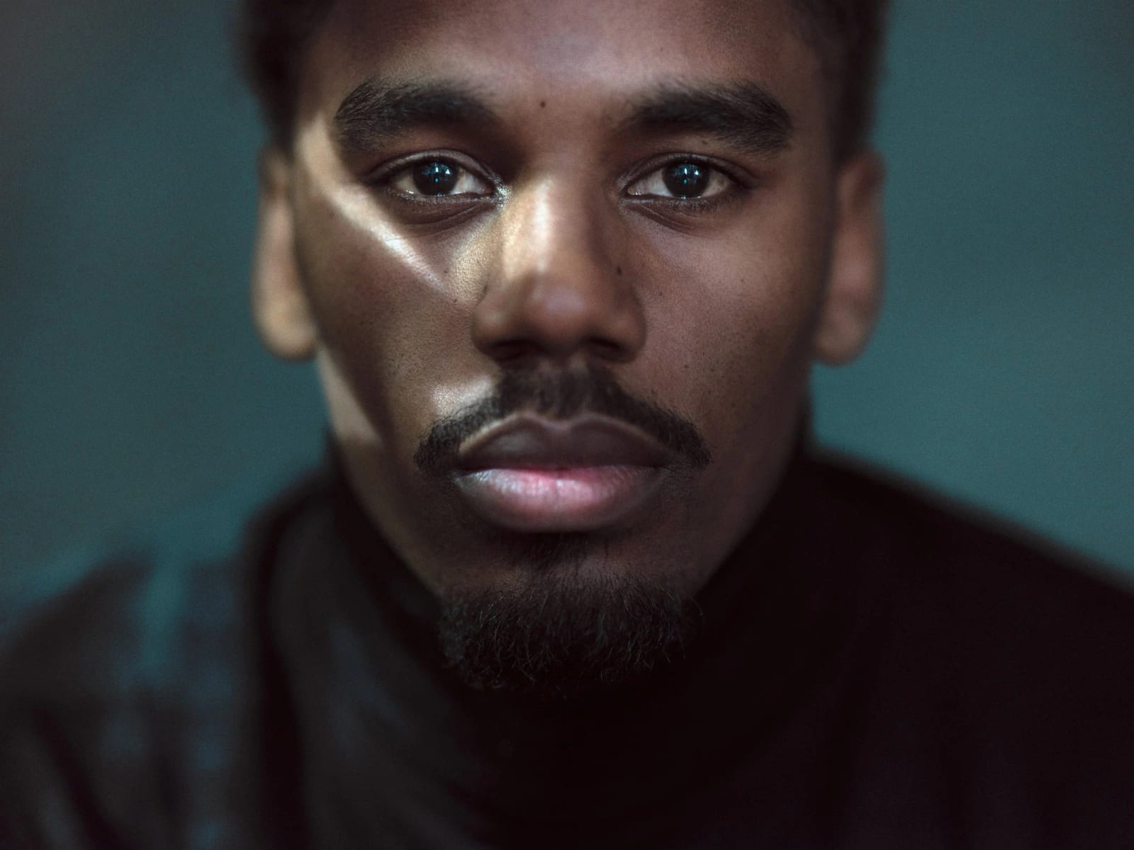 Close-up portrait of a man with a moustache and goatee in a black turtleneck, a band of light across his eyes.